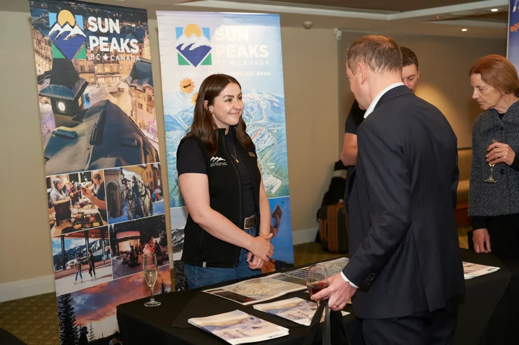 Tourism exhibition representative speaks with a suited attendee at a professional networking event in Melbourne, with Sun Peaks BC Canada promotional banners, travel brochures and alpine resort imagery displayed behind them.