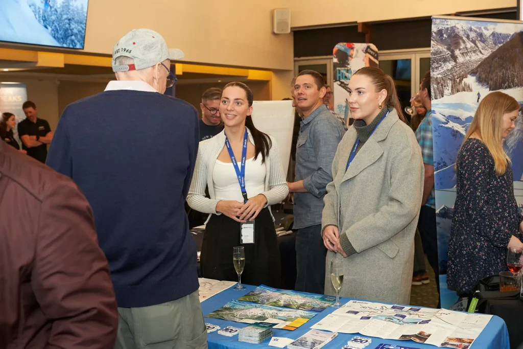 Travel exhibition representative speaks with attendees at a busy mountain resort trade show in Melbourne, with winter trail maps, brochures and ski resort banners surrounding the booth as visitors network and explore travel opportunities.