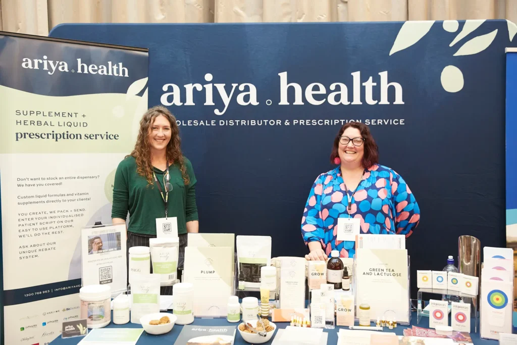 Two smiling wellness brand representatives pose behind a fully stocked exhibition booth in Melbourne, surrounded by natural health products, promotional materials and branded displays, capturing a friendly and professional team moment at a corporate trade show.