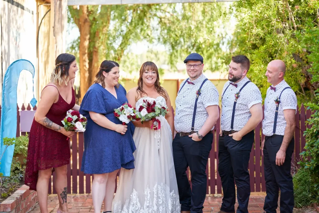 Bride and groom smiling at camera with bridesmaids and groomsmen looking at them, candid and relaxed with fence and green trees in background