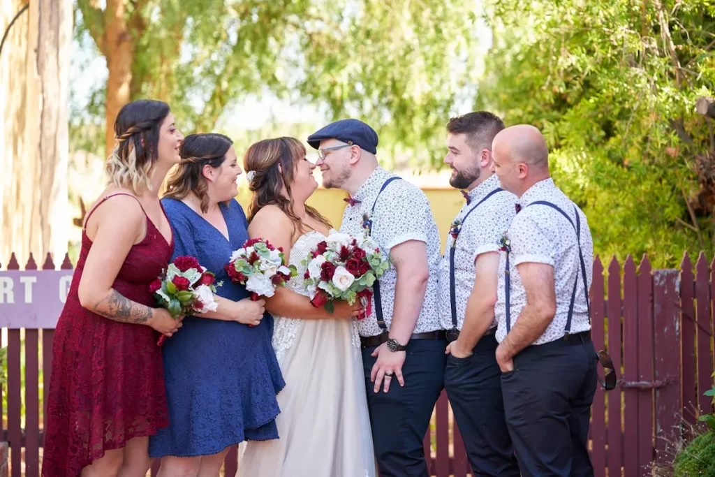 Bride and groom facing each other smiling before a kiss, with bridesmaids and groomsmen looking at them, candid and happy with fence and green trees in background