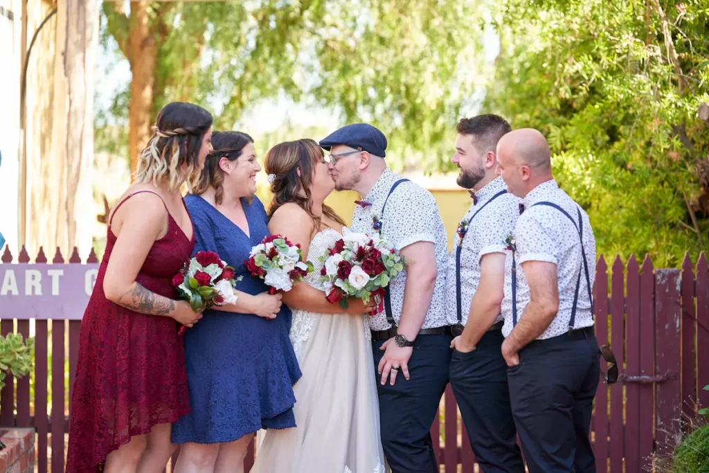 Bride and groom kissing with bridesmaids and groomsmen smiling and looking at them, candid and joyful with fence and green trees in background