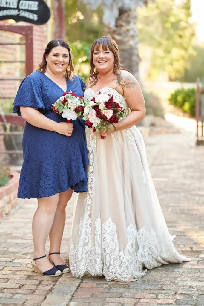 Bride and bridesmaids standing close, smiling and hugging with bouquets, creamy background of trees, plants, and golden light