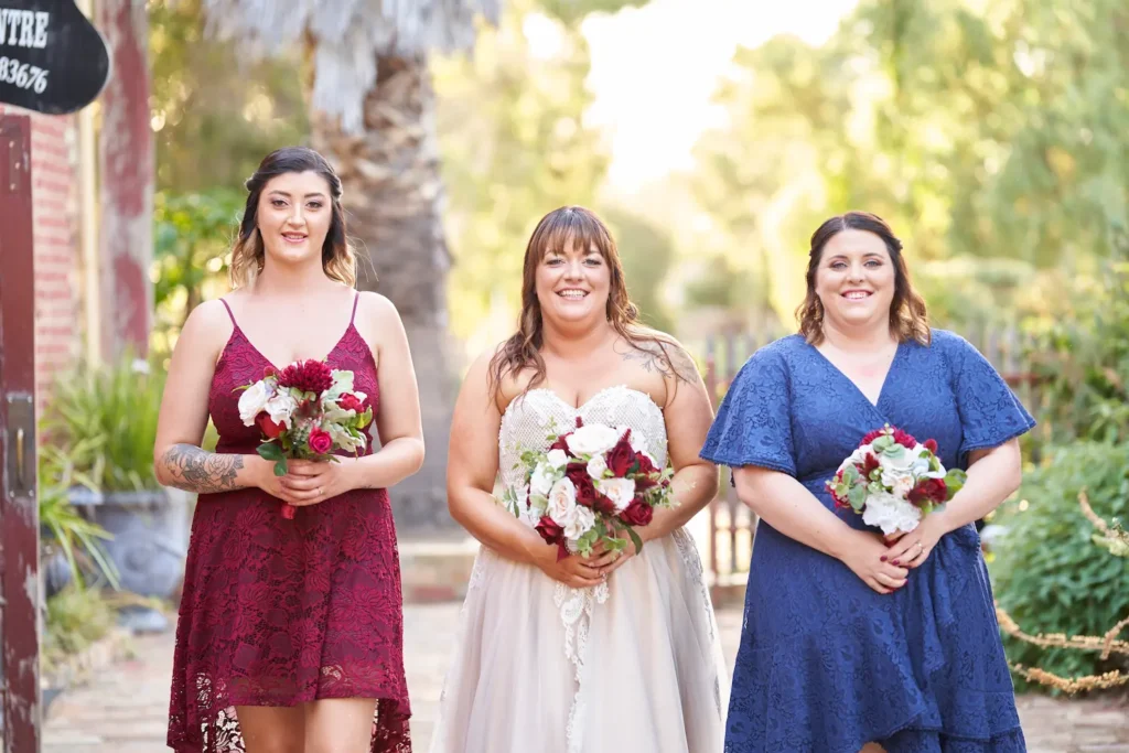 Bride and bridesmaids walking toward camera smiling, holding bouquets with creamy bokeh background of trees, plants, and golden light