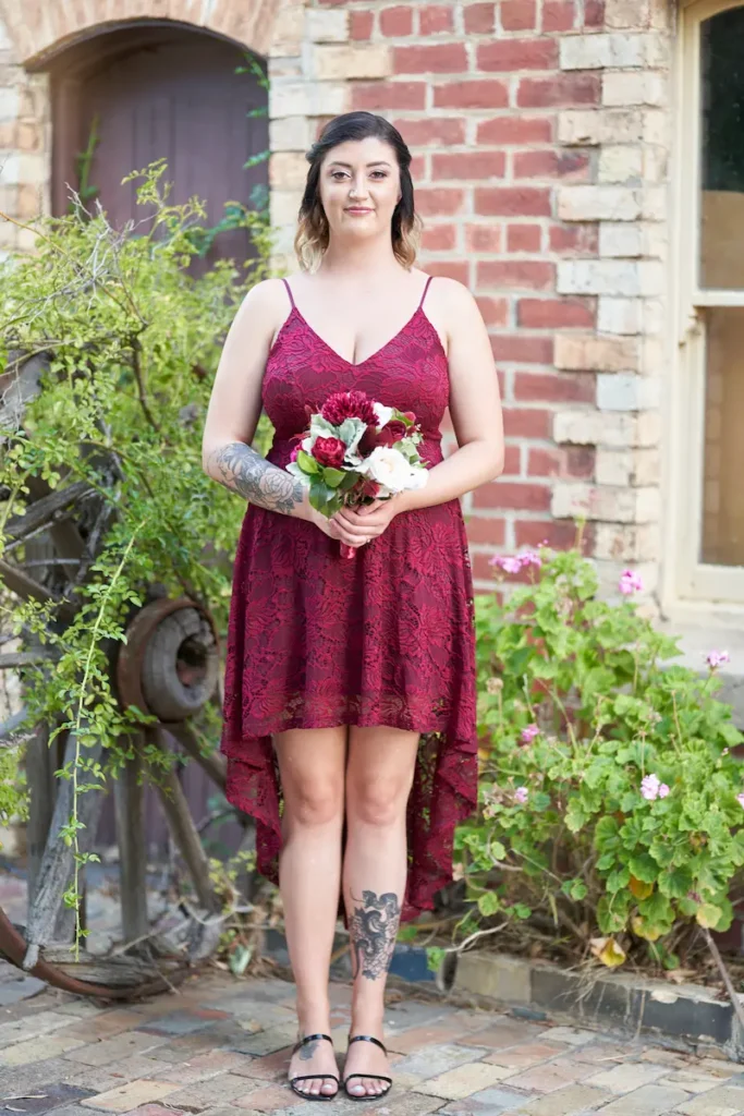 Bridesmaid in red dress smiling and holding bouquet with old wagon wheel, plants, and classic brick building in background