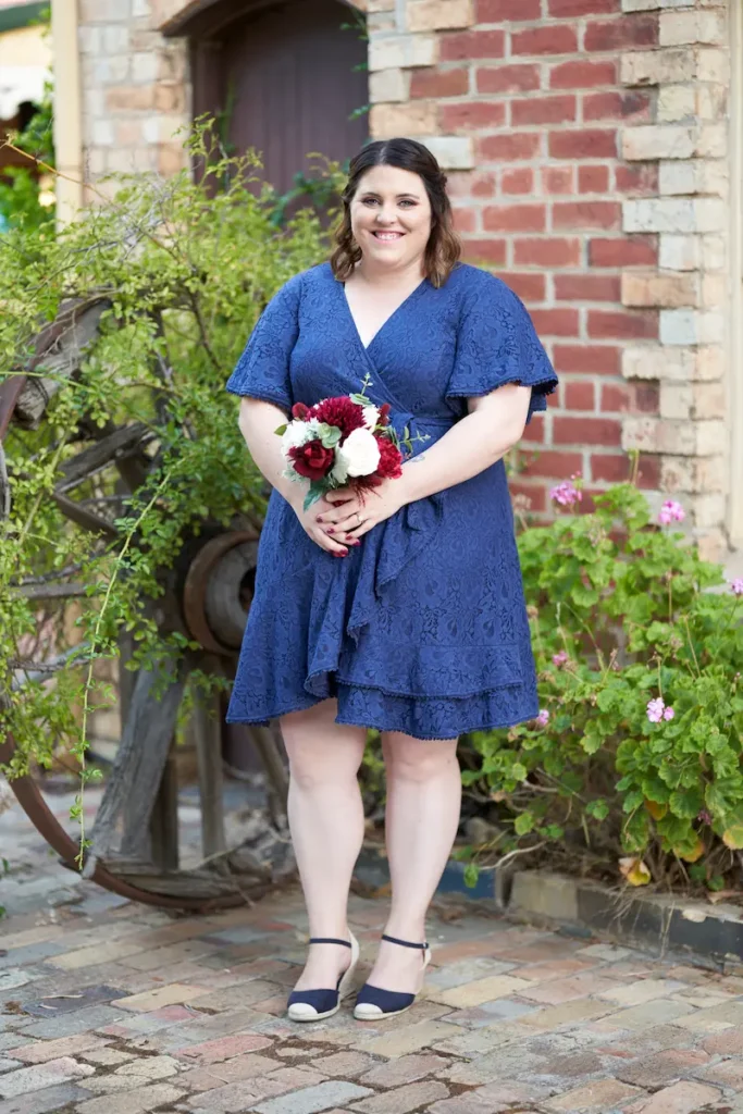 Bridesmaid in dark blue dress smiling and holding bouquet with old wagon wheel, plants, and classic brick building in background