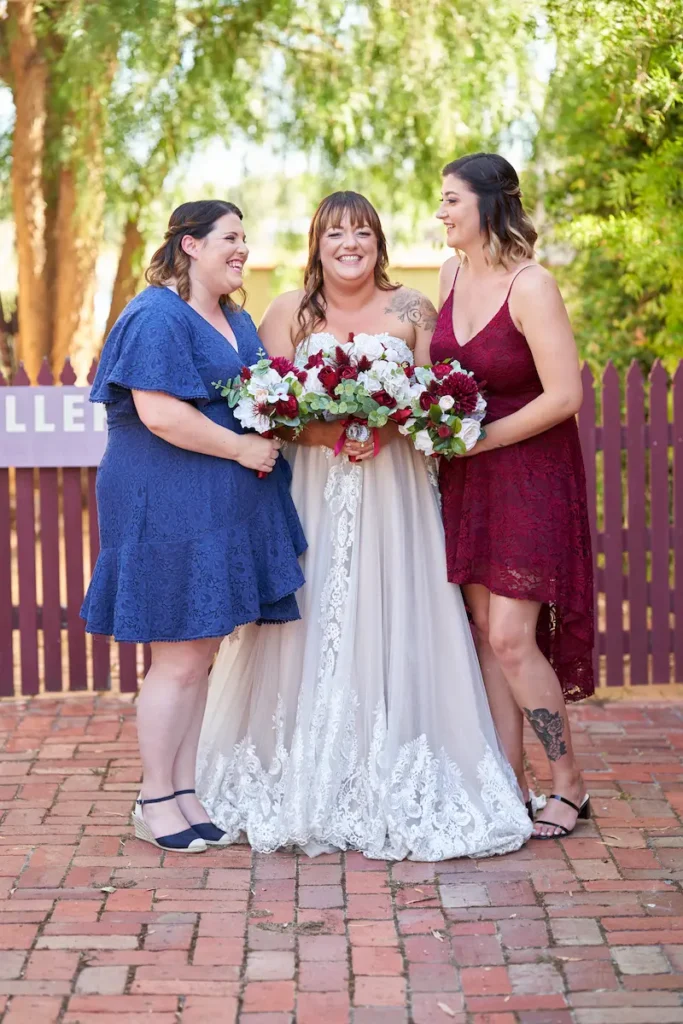 Bride smiling and laughing with bridesmaids looking at her, all holding bouquets with fence and green trees in background