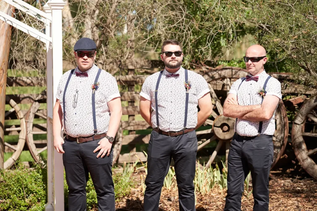 Groom and groomsman standing at wedding ceremony waiting for bride, sunny day with wooden fence, old wagons, and trees in background