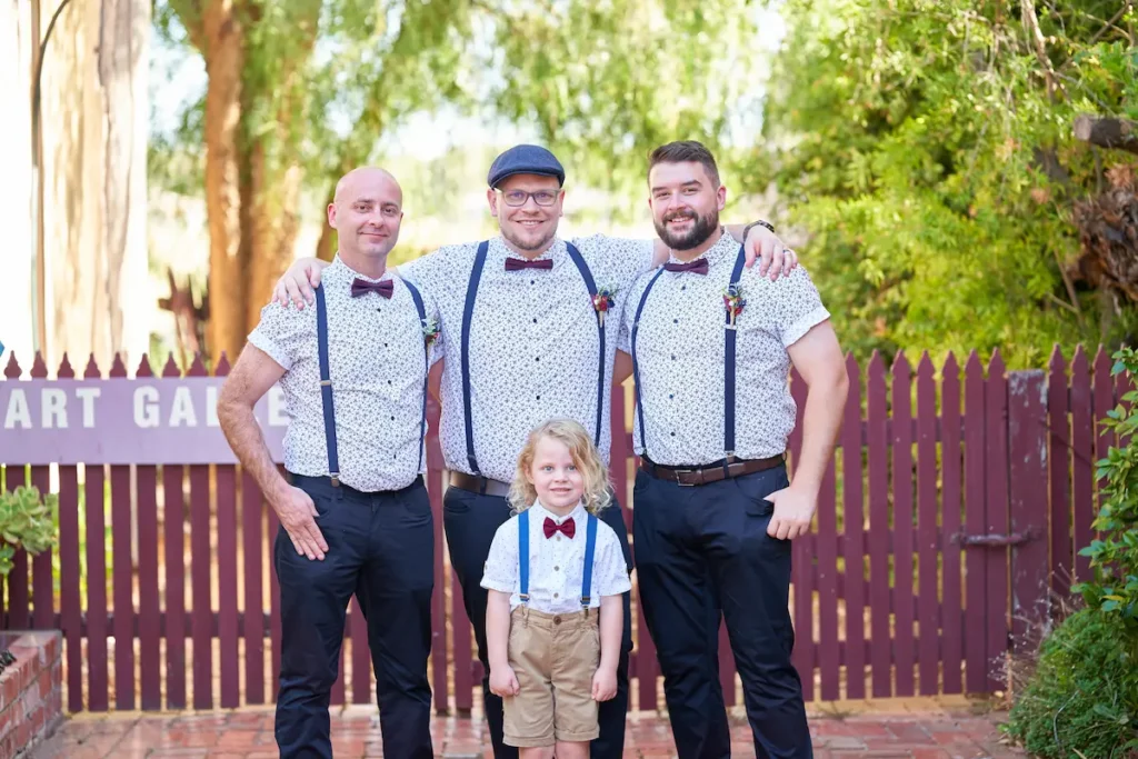 Groom with arm around groomsman’s shoulders, young boy in bow tie standing in front, all smiling with fence and green trees in background