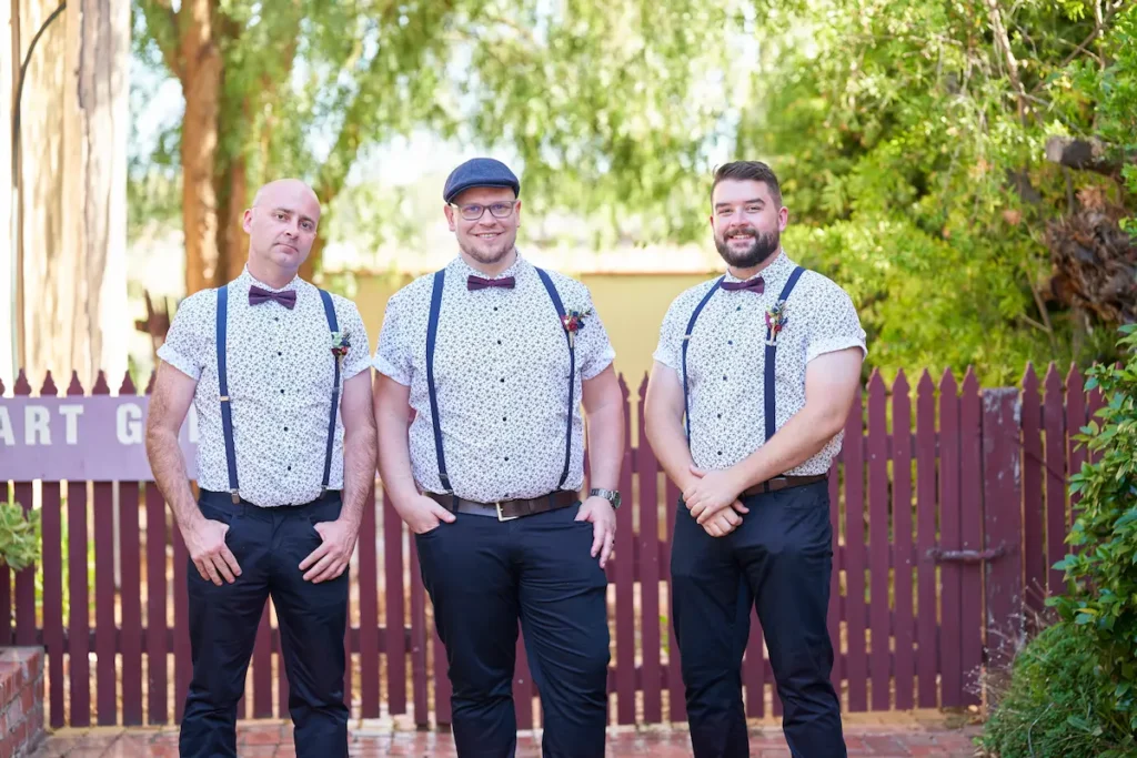 Groom and groomsman in casual happy portraits with fence and green trees in background