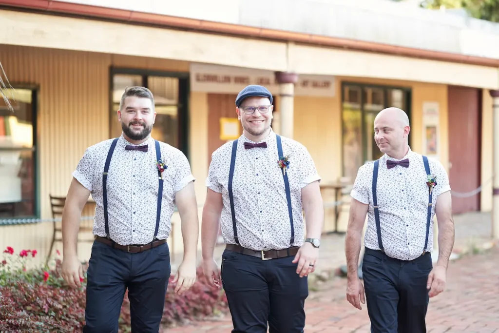 Groom and groomsman walking together smiling with soft light and red plants with small green and red flowers in background