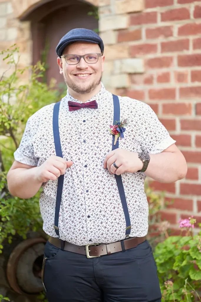 Groom standing and smiling with a friendly, happy pose against a background of green plants and brick wall