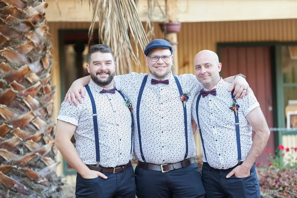 Groom with arm around groomsmen’s shoulders, all smiling in front of building with small flowers visible