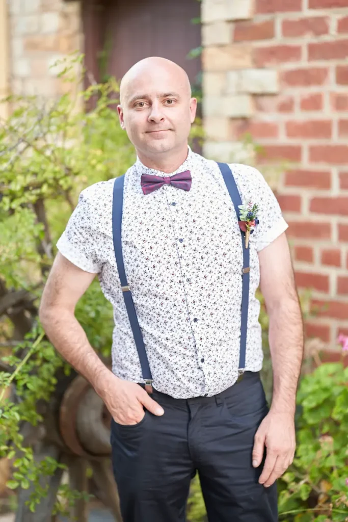 Groomsman standing and smiling with one hand in pocket, green plants and brick wall in the background