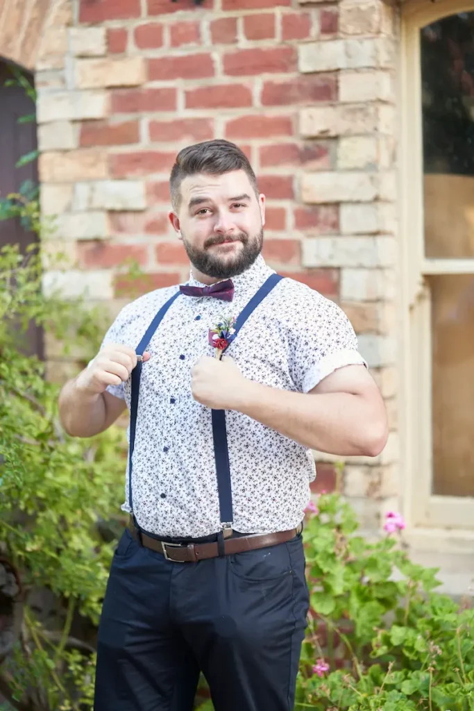 Groomsman in friendly casual pose with green plants and brick wall in the background