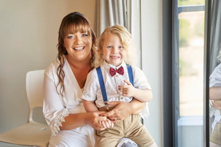 Bride sitting indoors with cute well-dressed boy wearing bow tie on her lap, both smiling in happy family moment during bride getting ready