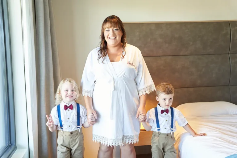 Bride standing indoors holding hands with two cute well-dressed little boys wearing bow ties during bride getting ready