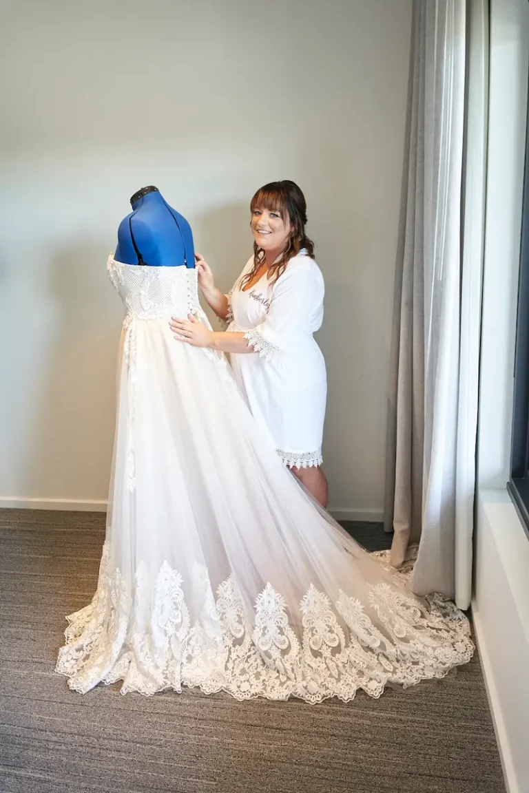 Bride standing by window with hand touching wedding dress before wearing it, soft light coming through window during bride getting ready