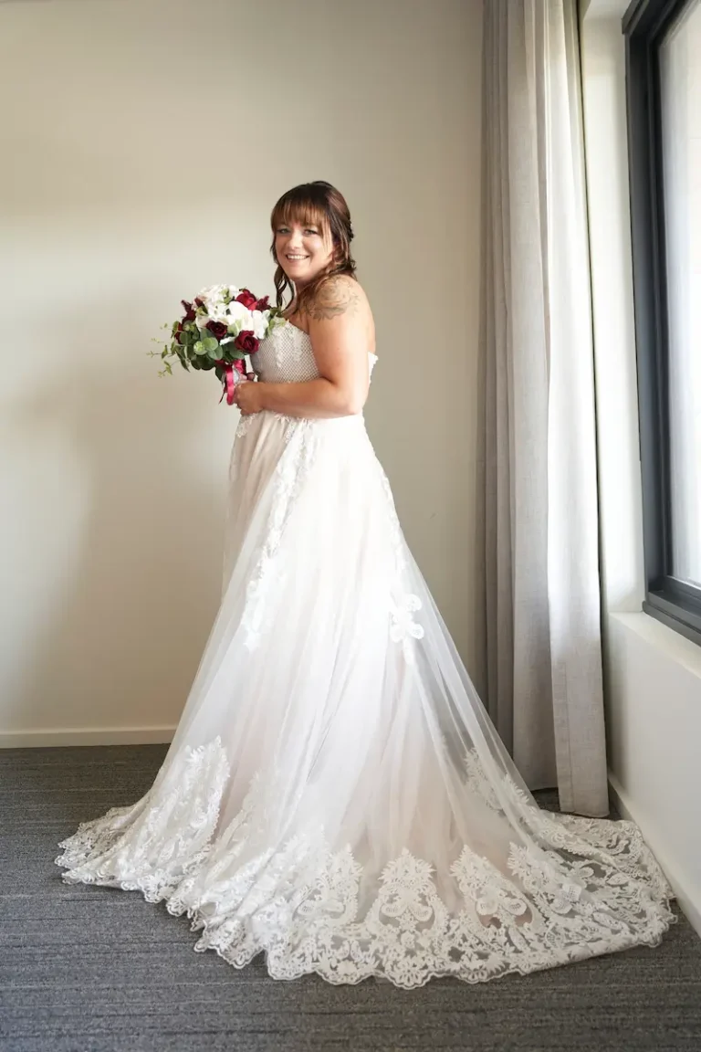 Portrait of bride standing sideways near large window with soft light, holding bouquet and smiling during bride getting ready
