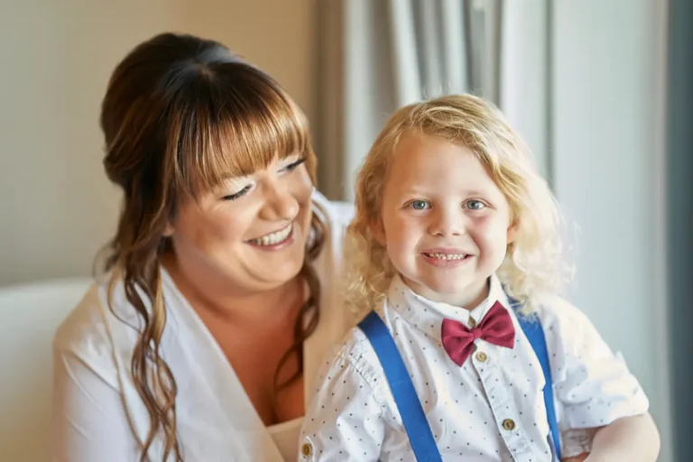 Close-up of bride sitting indoors looking at well-dressed boy wearing bow tie on her lap, both smiling in candid happy family moment during bride getting ready