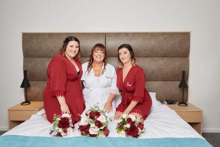 Bride and bridesmaids kneeling on bed smiling at camera, wearing bridal robes with bouquets on bed during bride getting ready