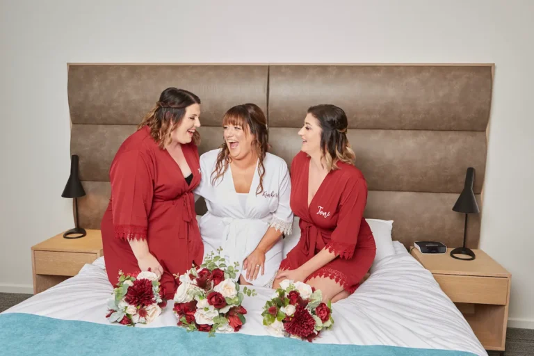 Bride and bridesmaids kneeling on bed laughing and looking at each other, wearing bridal robes with bouquets on bed during bride getting ready