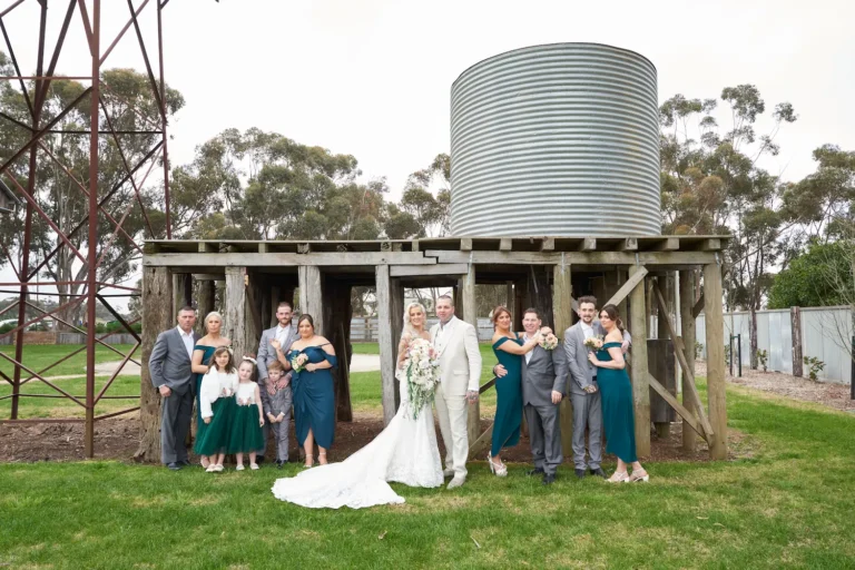 Group photo of bride Emma hugging groom Darko with one arm while holding her wedding bouquet in the other, smiling at the camera alongside bridesmaids, groomsmen, flower girls, and ring bearer. A rustic wooden structure with smaller silos sits in the background at Warrawong Estate