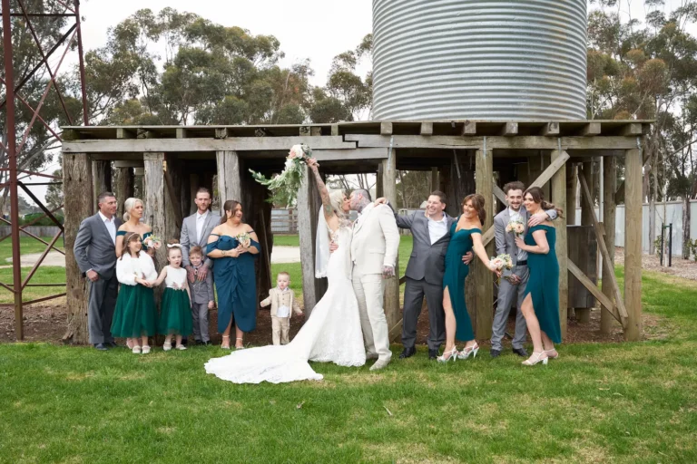 Group photo of bride Emma kissing groom Darko while hugging him with one arm and raising her wedding bouquet with the other. Bridesmaids, groomsmen, flower girls, ring bearer, and their young son join the joyful moment, with a rustic wooden structure and smaller silos in the background at Warrawong Estate