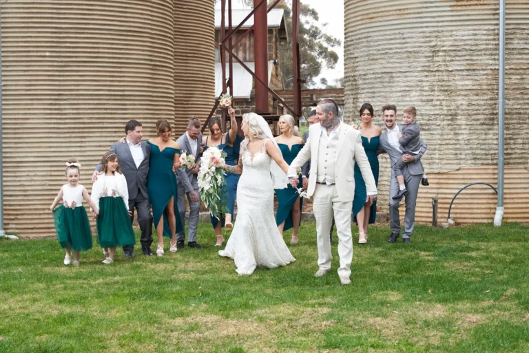 Candid group photo of bride Emma and groom Darko walking with bridesmaids, groomsmen, flower girls, and ring bearer at Warrawong Estate. Emma looks back at her bridesmaids with laughter, creating a joyful moment, with the estate’s massive silos in the background