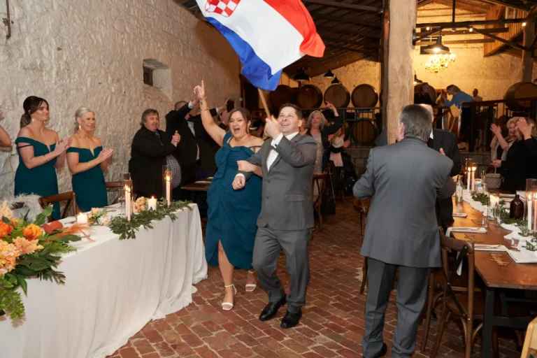 Bridesmaids and groomsmen entering the wedding reception at Warrawong Estate, holding hands while a groomsman carries a flag. Guests stand and clap joyfully to welcome them