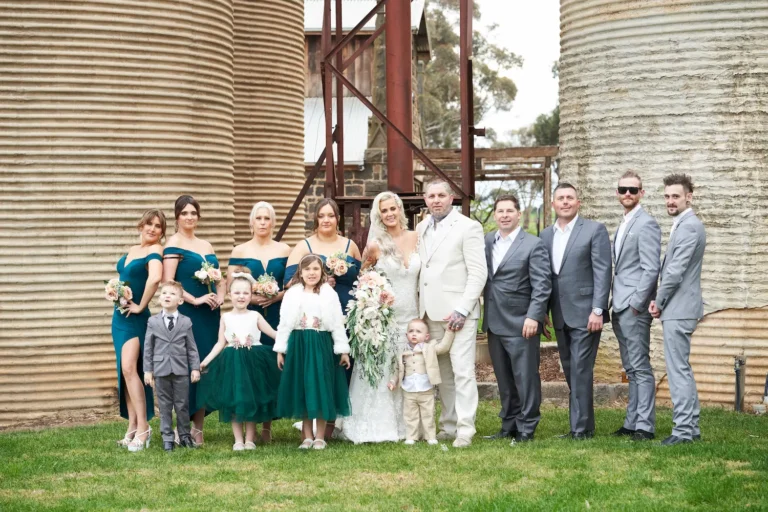 Formal group photo of bride Emma and groom Darko with bridesmaids, groomsmen, flower girls, ring bearer, and their young son at Warrawong Estate. The estate’s massive silos, a unique landmark, stand prominently in the background