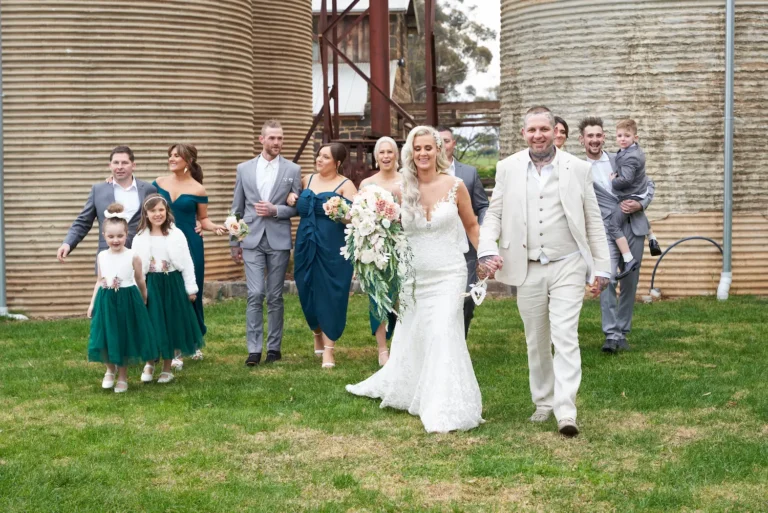 Group photo of bride Emma and groom Darko holding hands while walking toward the camera with bridesmaids, groomsmen, flower girls, and ring bearer at Warrawong Estate. The estate’s massive silos, a unique landmark, rise in the background