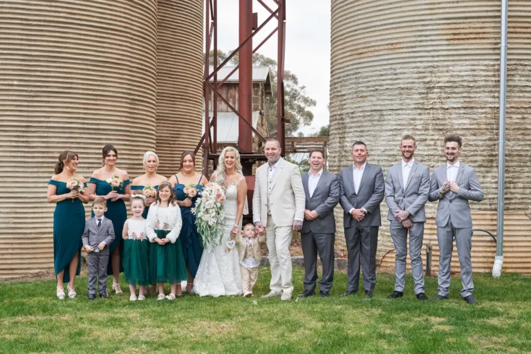 Relaxed group photo of bride Emma and groom Darko with bridesmaids, groomsmen, flower girls, ring bearer, and their young son at Warrawong Estate. Everyone smiles and laughs in a joyful moment, with the estate’s massive silos as the backdrop