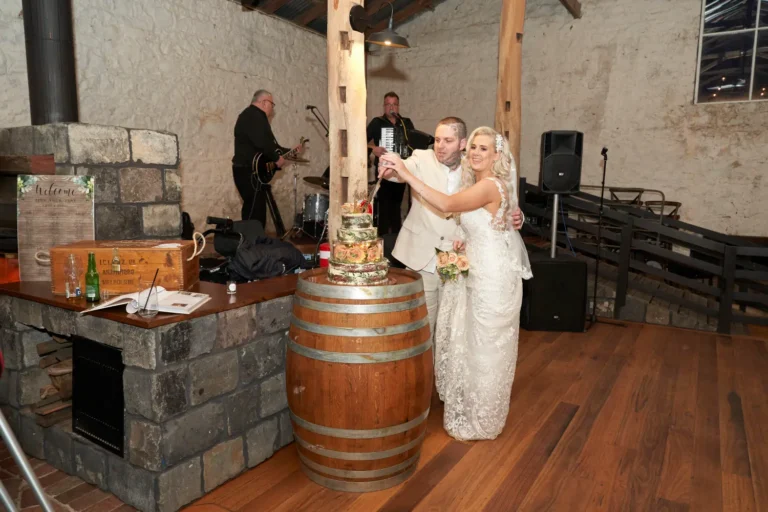 Bride Emma and groom Darko holding hands while cutting their wedding cake, which is placed on top of a wine barrel, at Warrawong Estate. A musician plays in the background, adding to the celebratory atmosphere