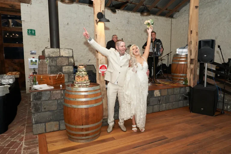 Bride Emma and groom Darko hugging and waving their hands in the air in celebration after cutting their wedding cake at Warrawong Estate, with a musician playing in the background