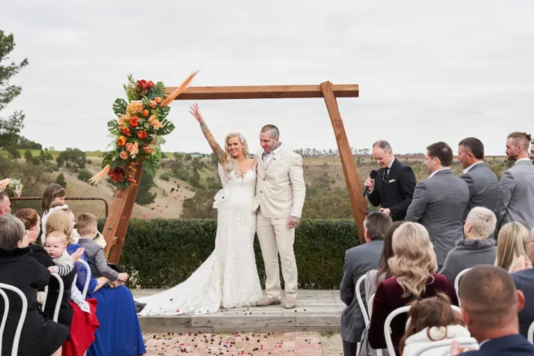 Bride Emma and groom Darko hugging after their Warrawong Estate wedding ceremony, as Emma waves to the guests, with the wedding arch framing the scenic landscape and valley under overcast skies