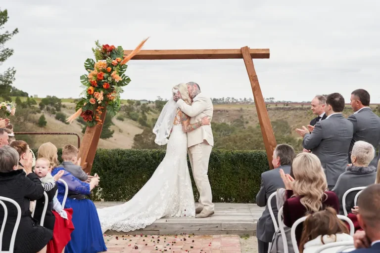 Bride Emma and groom Darko sharing their first kiss as husband and wife at Warrawong Estate, with guests clapping, the wedding arch framing them, and the scenic landscape and valley visible under overcast skies