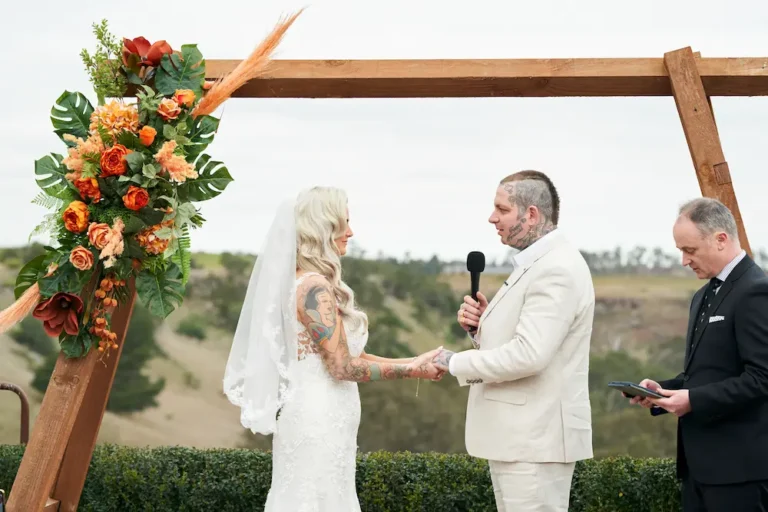 Groom Darko holding a microphone and speaking during Emma and Darko’s Warrawong Estate wedding ceremony, while they hold hands and face each other, with the wedding arch, scenic landscape, and valley visible under overcast skies