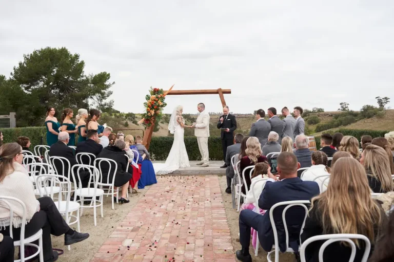 Wide shot of bride Emma and groom Darko holding hands during their Warrawong Estate wedding ceremony, with bridesmaids and groomsmen watching, scenic hills, open grass, a distant large tree, and the valley visible under overcast skies