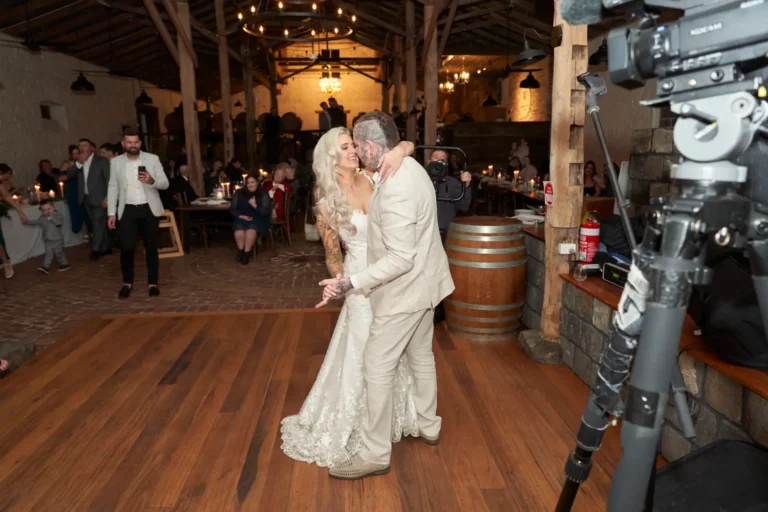 Wide shot of bride Emma and groom Darko holding hands and looking at each other lovingly during their first dance at Warrawong Estate. Guests watch from tables, some filming the moment, with the reception room softly illuminated by chandeliers