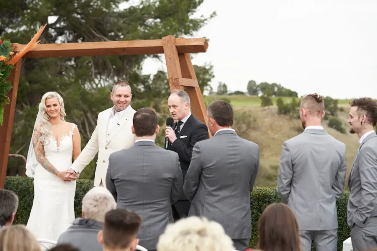 Bride Emma holding groom Darko’s hands and smiling during their Warrawong Estate wedding ceremony, with four groomsmen facing them, a large tree, and scenic landscape in the background