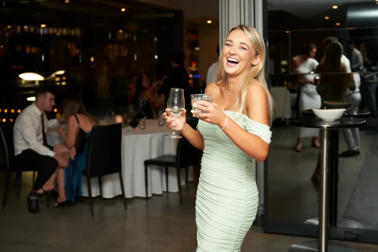 A smiling blonde woman in a light green off-the-shoulder dress holds a glass of white wine and a glass of water, laughing at the Garryowen Awards Night at River’s Edge Events, with other guests and tables visible in the dimly lit background