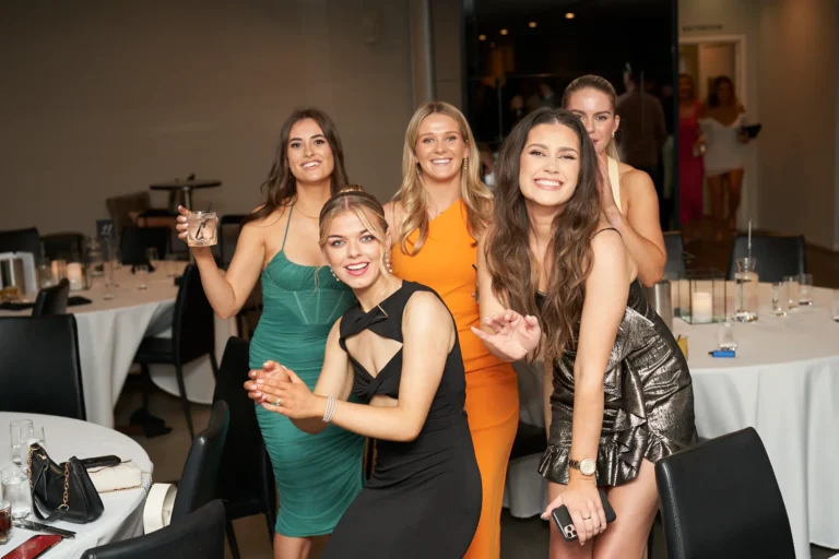 A group of five smiling young women in formal dresses posing together at the Garryowen Awards Night at River’s Edge Events with tables and chairs visible in the background. Three women are in the foreground: one in a green dress holding a drink, one in a black dress with her hands clasped, and one in an orange dress. Another woman in a black and gold patterned dress is slightly behind them, and another woman is visible further back
