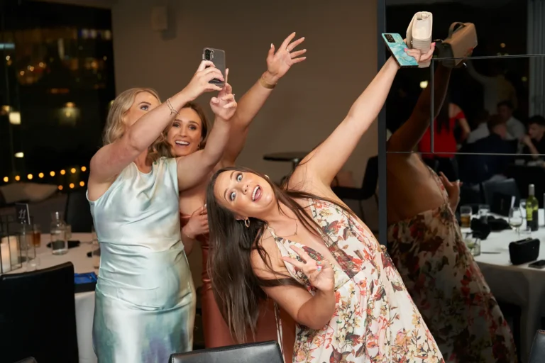 Three women in dresses taking selfies and posing at the Garryowen Awards Night at River’s Edge Events, with one holding a phone high and another making a peace sign with a playful pose
