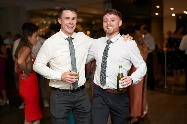 garryowen awards night rivers edge candid two men drinks Two smiling men in collared shirts and patterned ties stand with arms around each other at the Garryowen Awards Night at River’s Edge Events, one holding a glass and the other a green bottle, with other guests blurred in the background
