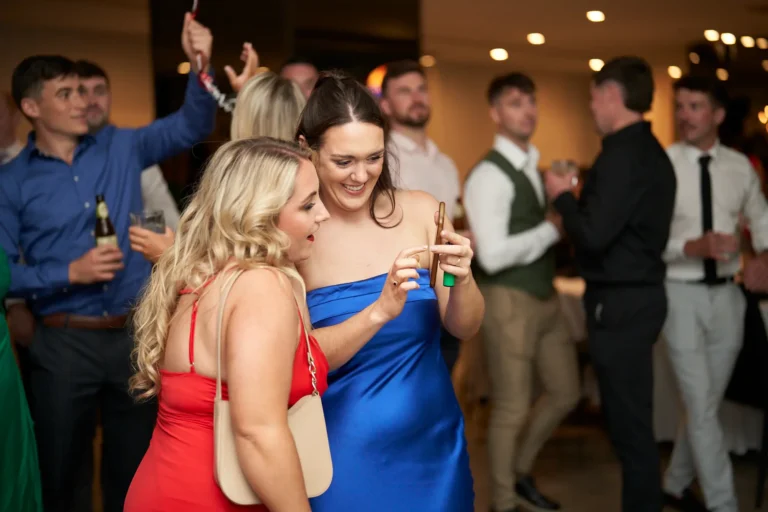 Two women in formal attire, one in a red dress and another in a blue strapless dress, looking at a phone together at the Garryowen Awards Night at River’s Edge Events, with other attendees visible in the background