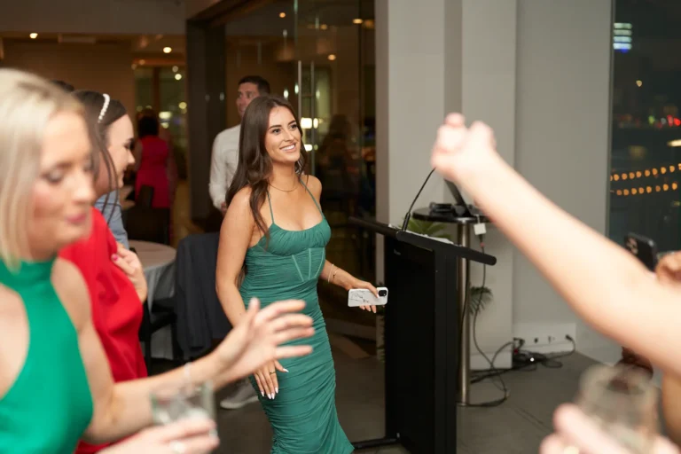 A young woman in a teal green ruched slip dress smiles near a black podium at the Garryowen Awards Night at River’s Edge Events, holding a phone while looking to the left, with other guests blurred in the foreground and background and city views through large windows at night