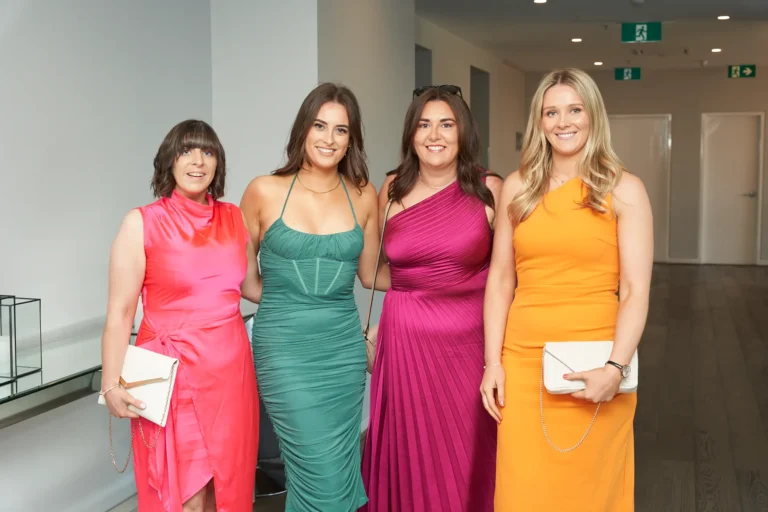 Four women in vibrant formal dresses posing together while arriving at River’s Edge Events for the Garryowen Awards Night. The woman on the left wears a pink dress, the second from left wears a green dress, the third wears a fuchsia dress, and the woman on the right wears an orange dress. They are smiling and holding clutch bags