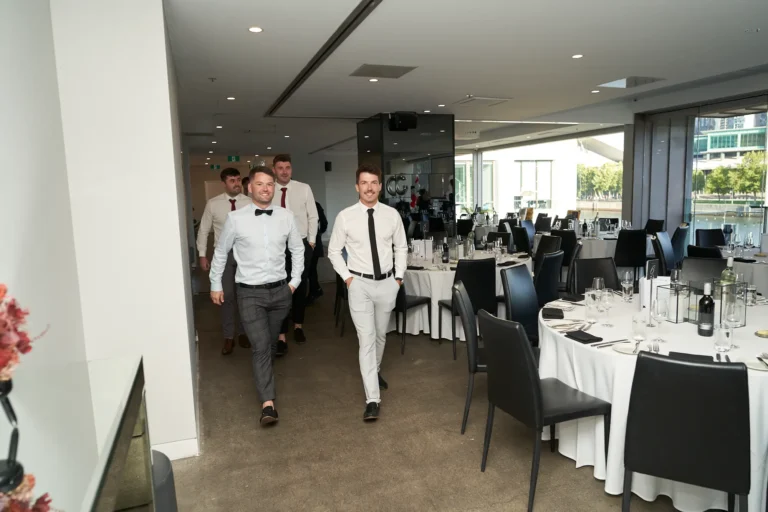 Group of men in formal attire walking down a hallway into the event room at River’s Edge Events for the Garryowen Awards Night, with round tables and waterfront views through large windows
