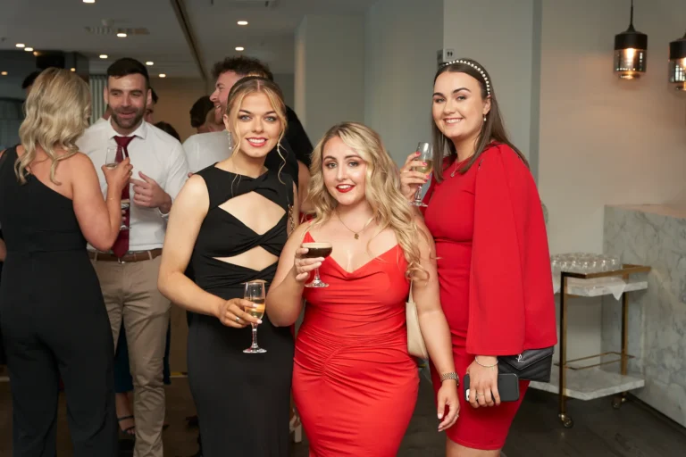 Three women smiling and holding drinks at River’s Edge Events during the Garryowen Awards Night, with other attendees and a bar softly lit in the background